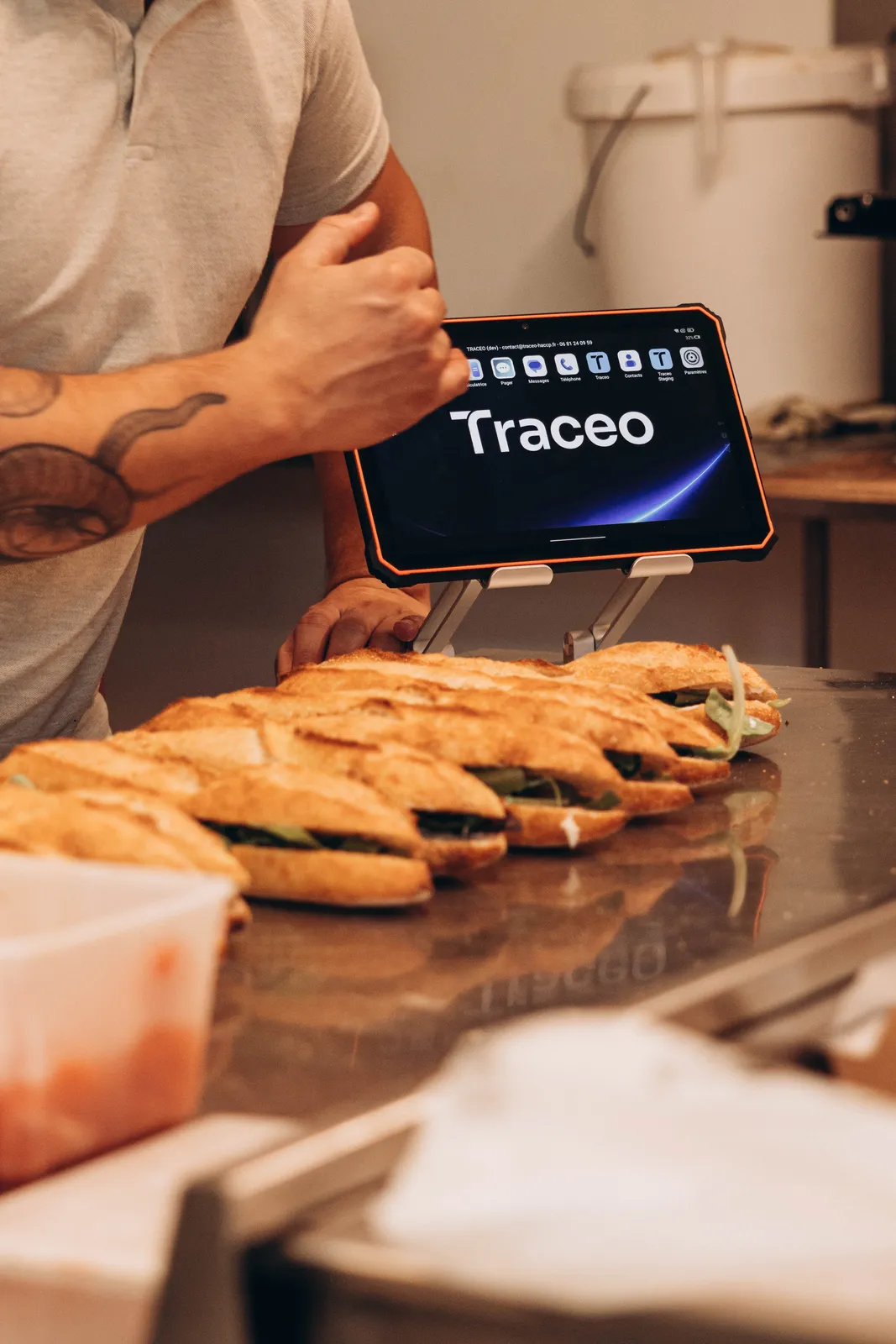 Traceo-nettbrett på et stativ bak en rekke smørbrød som tilberedes i et bakeri.